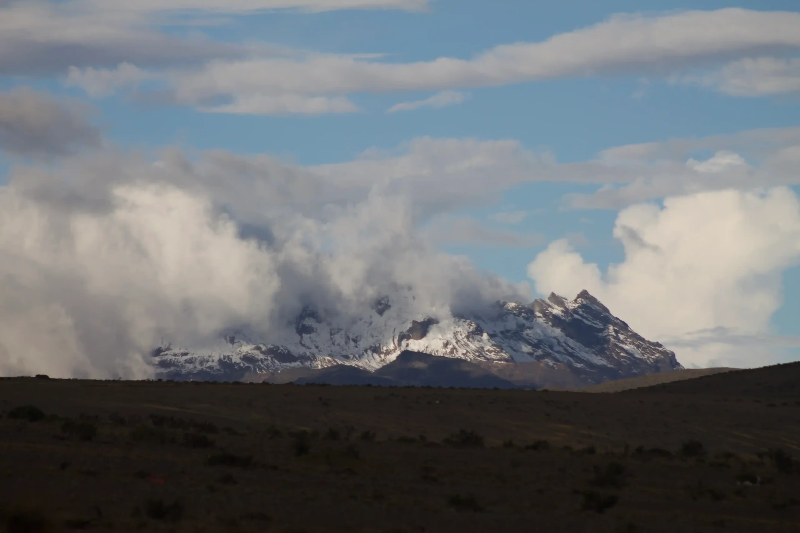 Imponente montaña nevada con picos escarpados parcialmente envuelta en densas nubes blancas, vista desde la llanura oscura de un páramo andino.